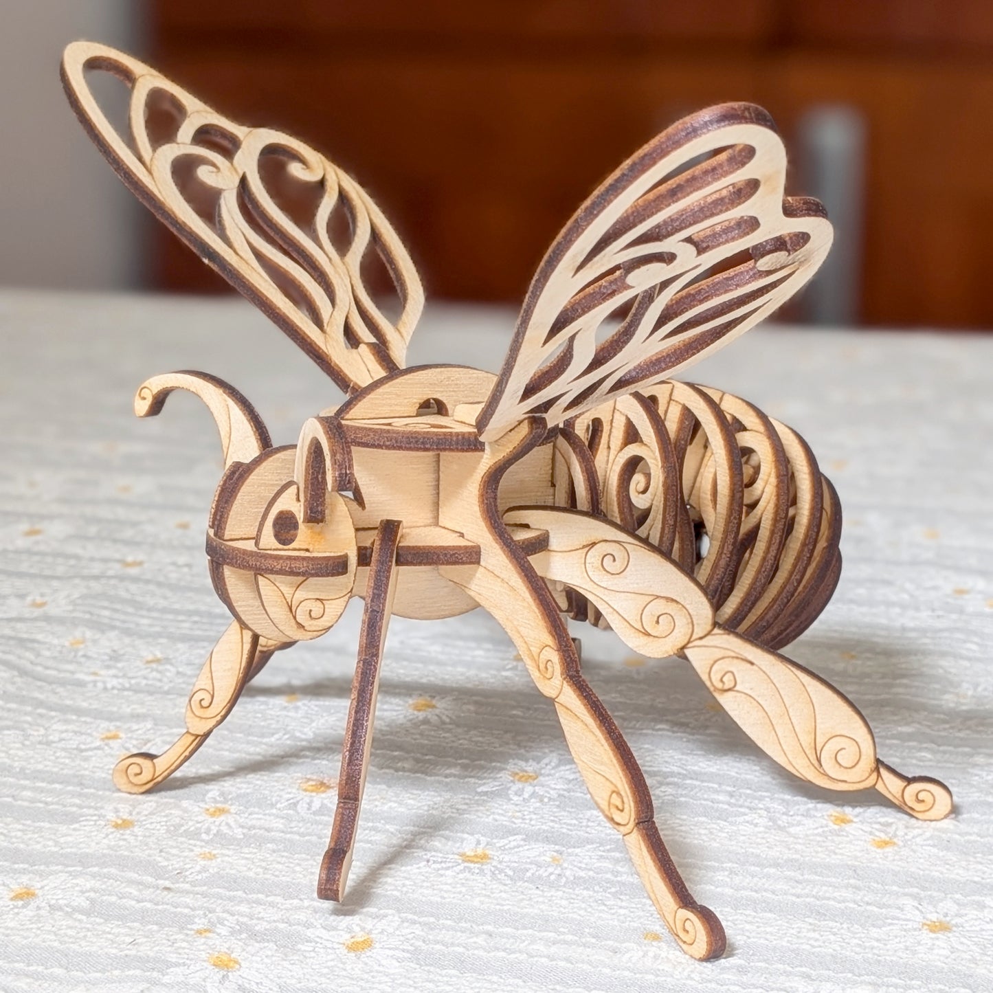 Laser cut wooden fairy bee ornament with elegant layered plywood design, featuring intricate wings and detailed scroll patterns, displayed on a light textured tablecloth