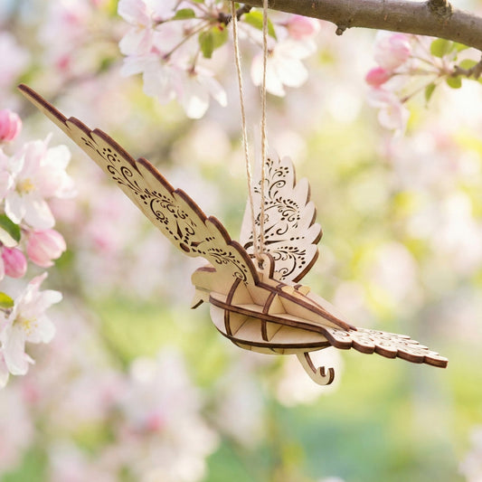 Back view of a hanging fairy bird decoration crafted from laser cut plywood. Detailed wood-burned patterns decorate the wings and tail. It is suspended from a tree branch surrounded by blooming spring flowers in a soft-focus garden.