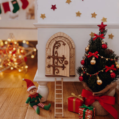 Laser cut plywood fairy door set with ladder and elf figurine staged near miniature Christmas tree, gifts, and glowing lights.