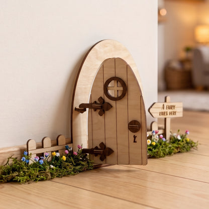 Open laser cut plywood fairy door set with picket fence, moss, tiny flowers, and sign against white baseboard and wood floor.