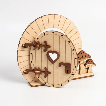 Round hobbit style laser cut plywood fairy door with heart window, decorative strap hinges, and little mushrooms on white base.