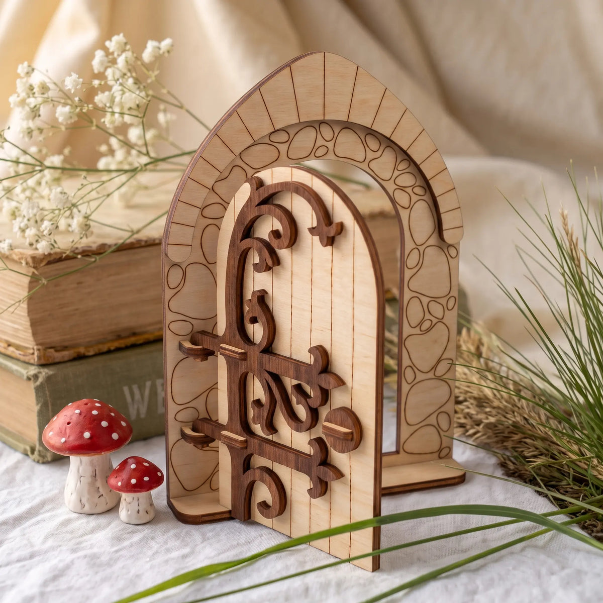 Medieval style arched laser cut plywood fairy door model staged on white table beside old vintage books and white flowers.