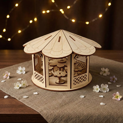 Miniature gazebo made of laser cut plywood with a scalloped roof and lattice walls. Small flower-shaped stools are visible inside. The piece is surrounded by white spring petals on a linen surface with soft bokeh lighting.