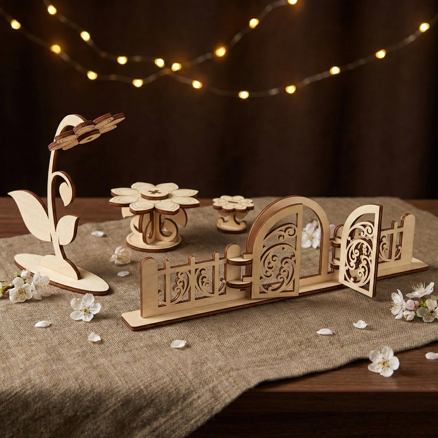 Ornate garden fence and open gate made of laser cut plywood with delicate scrollwork patterns. A tall wooden flower and tables stand in the background. The scene is illuminated by warm fairy lights for a magical atmosphere.