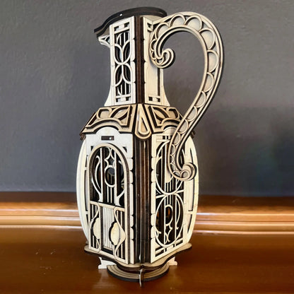 Ornate laser cut plywood jug fairy house model standing on a polished wooden surface against a dark grey background wall.