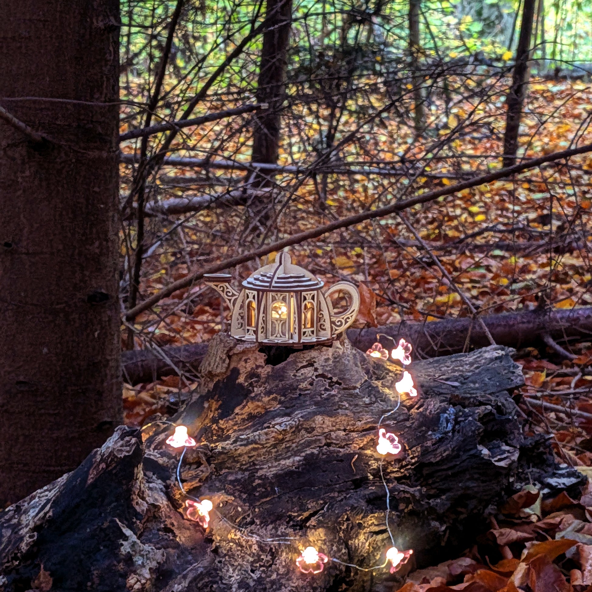 Wooden laser cut fairy kettle house lantern placed on a log in an autumn forest, surrounded by soft glowing flower lights