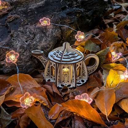 Fairy teapot house lantern made from laser cut plywood surrounded by glowing flower lights and autumn leaves
