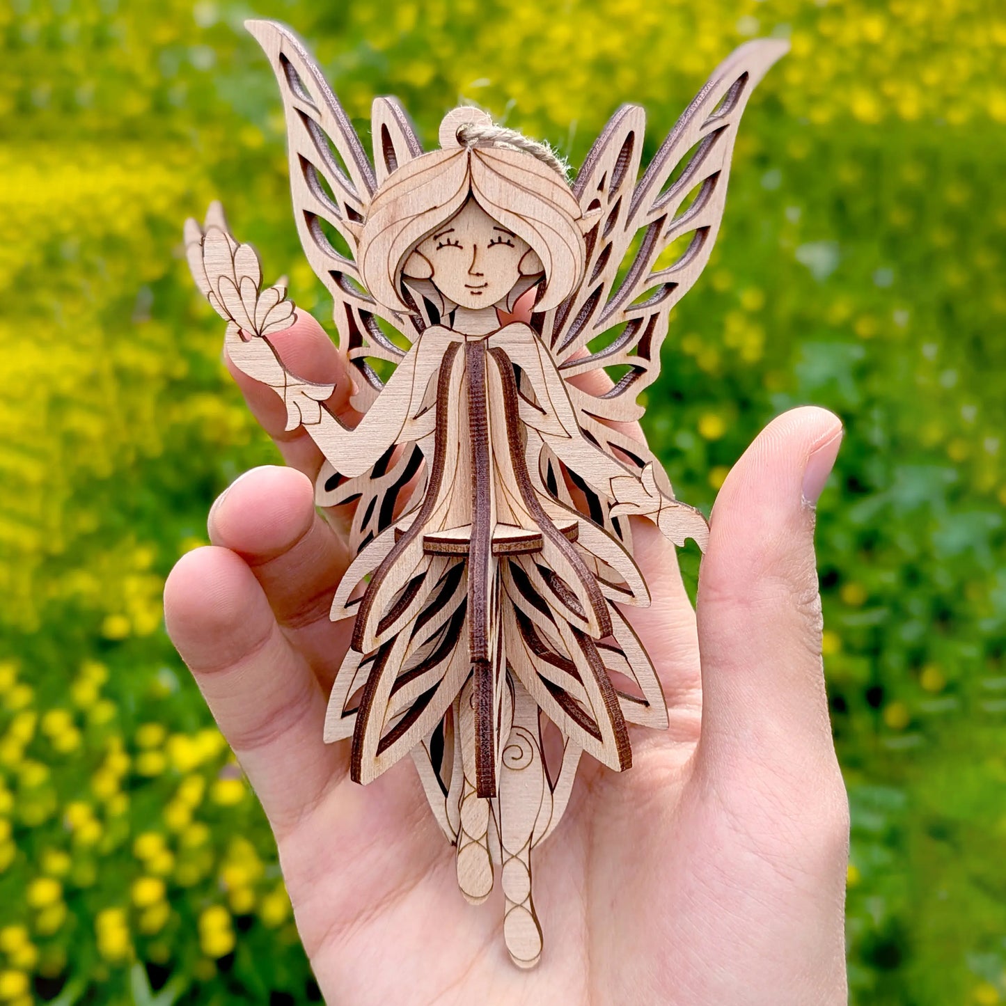 Wooden laser cut fairy ornament held in a hand with a blurred green and yellow background
