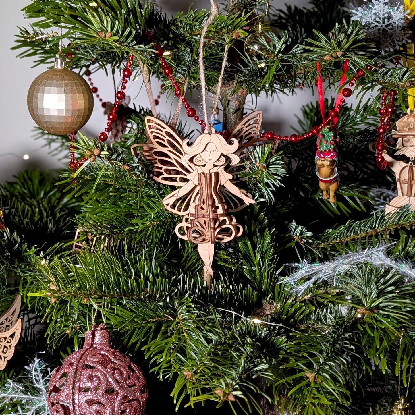 A 3D laser cut plywood fairy ornament hanging on a festive Christmas tree decorated with red beads and golden ornaments.