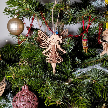 A 3D laser cut plywood fairy ornament hanging on a festive Christmas tree decorated with red beads and golden ornaments.
