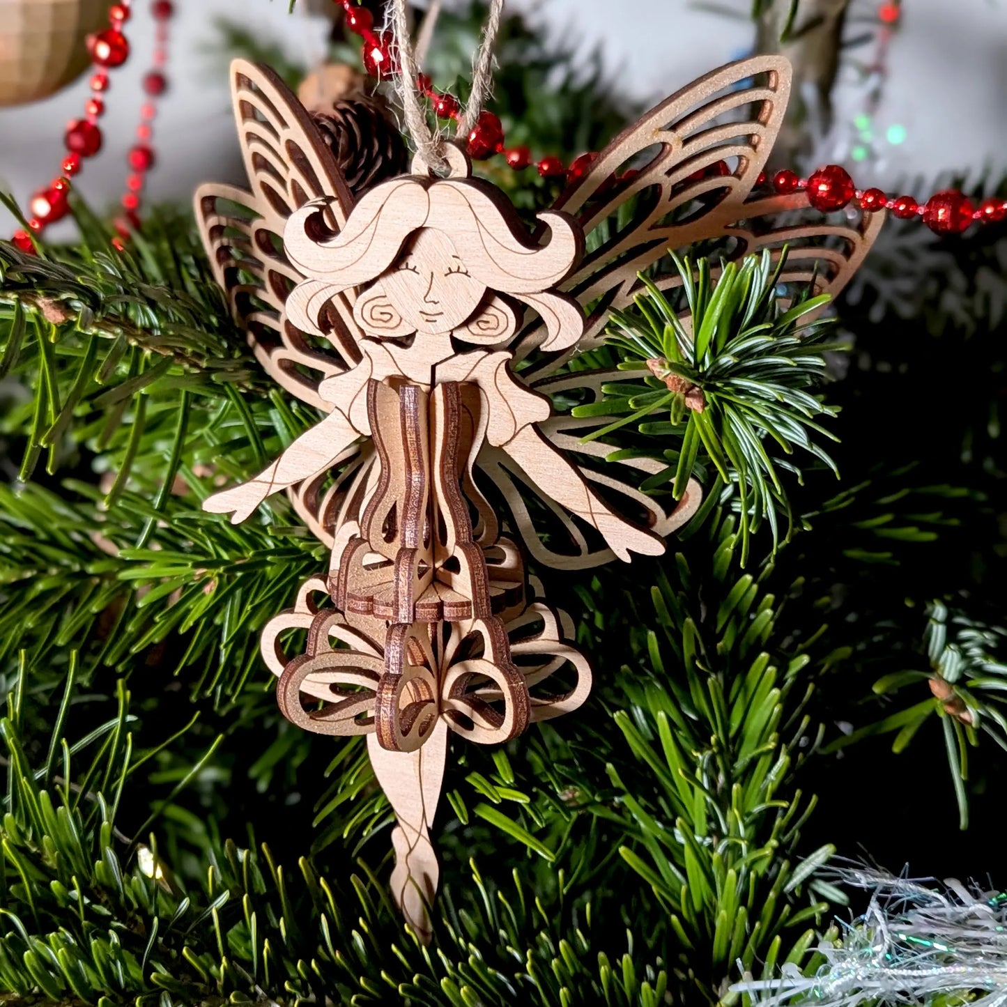 A 3D laser cut plywood fairy ornament displayed on a table with a red gift, gold baubles, and natural pinecone decor