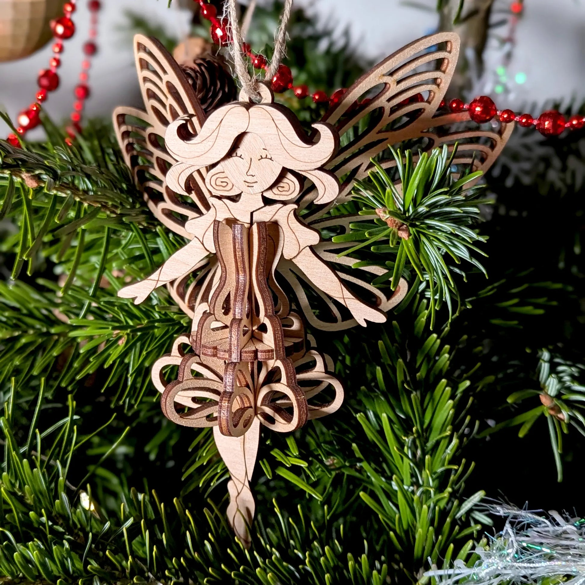 A 3D laser cut plywood fairy ornament displayed on a table with a red gift, gold baubles, and natural pinecone decor