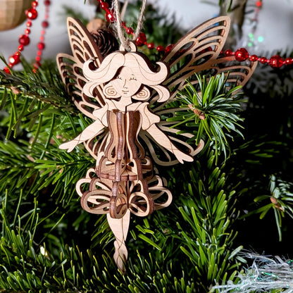 A 3D laser cut plywood fairy ornament displayed on a table with a red gift, gold baubles, and natural pinecone decor