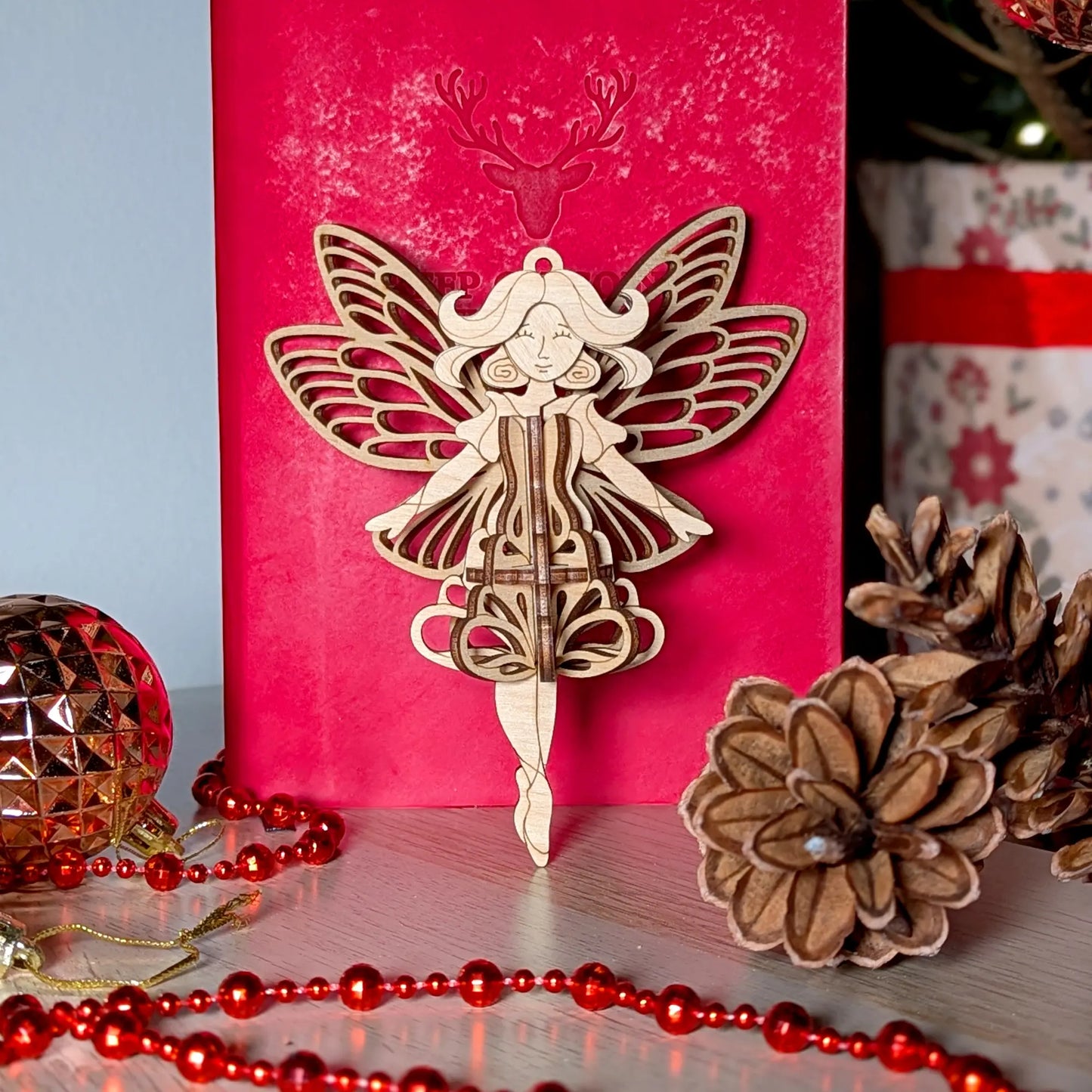 Elegant 3D laser cut plywood fairy ornament set against a red gift box with pinecones and festive gold bead decoration.
