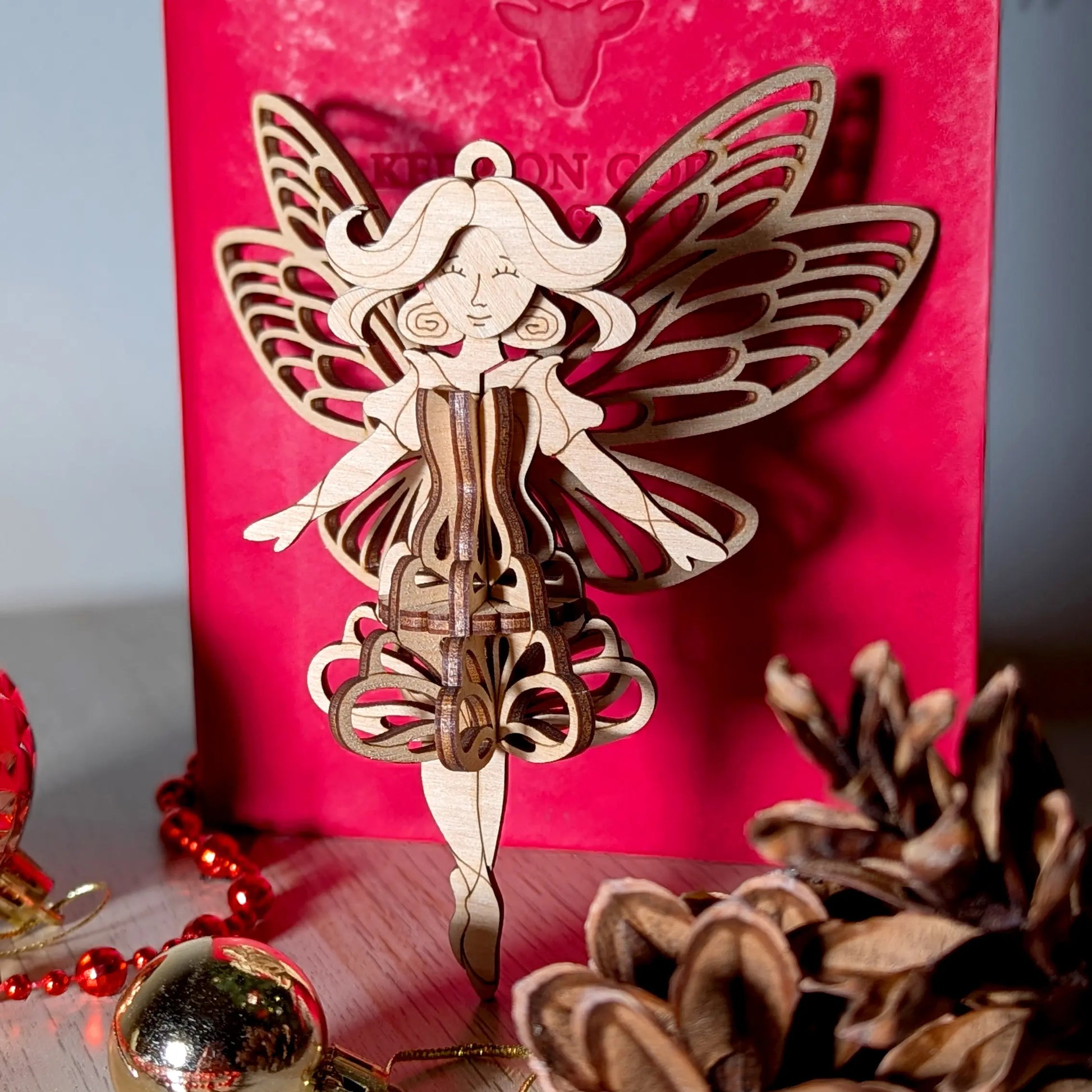 3D laser cut plywood fairy ornament placed on a festive holiday table with gold baubles, red beads, and pinecone decor.