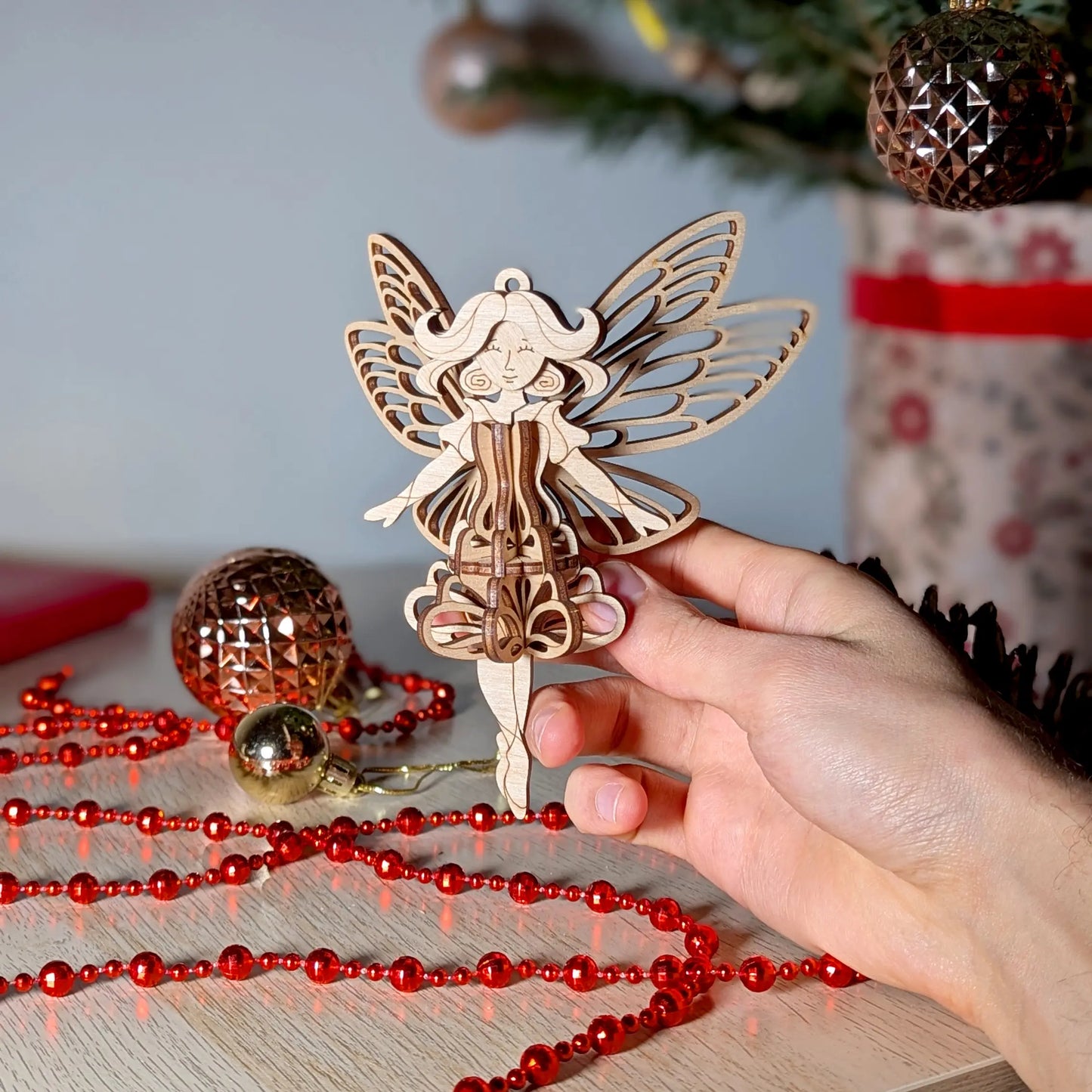 A hand holding a detailed 3D laser cut plywood fairy ornament with butterfly wings over a desk with holiday decorations.