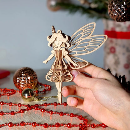 Person holding a 3D laser cut plywood fairy ornament with ornate wings against a festive table with red holiday garlands.