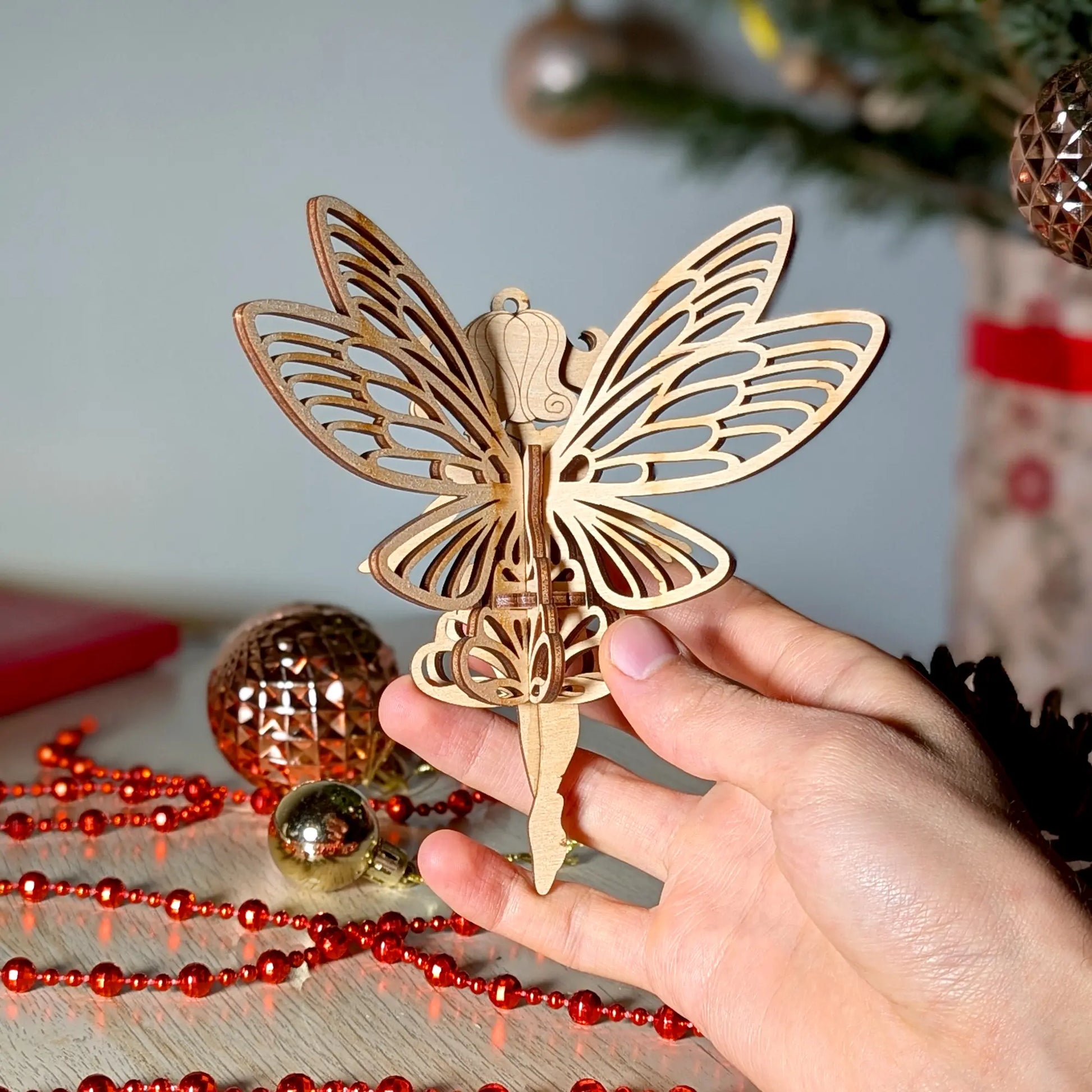 Person holding a 3D laser cut plywood fairy ornament with ornate wings against a festive table with red holiday garlands.
