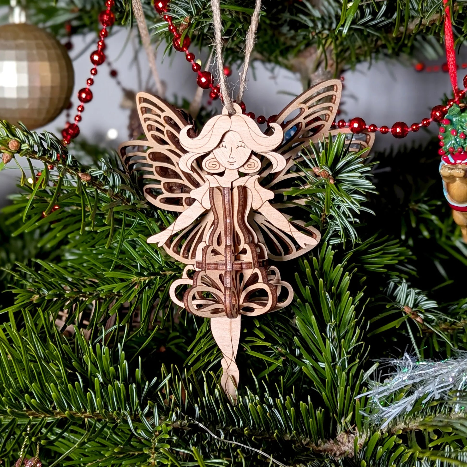 Close-up of a 3D laser cut plywood fairy ornament with ornate wings hanging on a lush green Christmas tree with lighting.