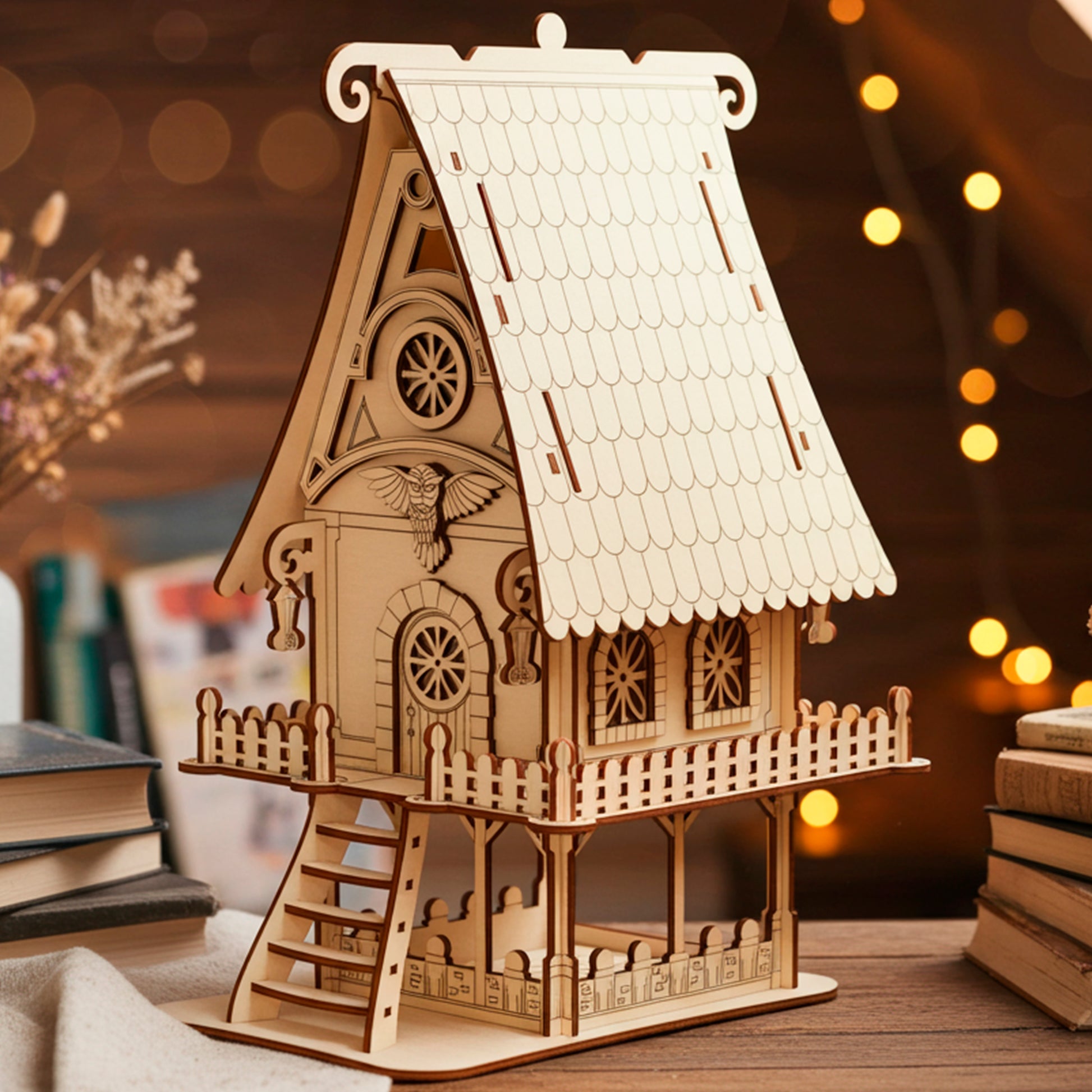 Laser cut  plywood detailed wooden fairy house or owl cottage model with a shingled roof, bird carving, and front balcony. The miniature house is placed on a wooden attic table surrounded by stacked vintage books and warm fairy lights.