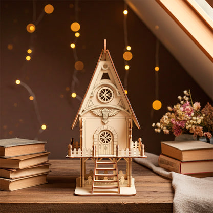 Laser cut  plywood detailed wooden fairy house or owl cottage model with a shingled roof, bird carving, and front balcony. The miniature house is placed on a wooden attic table surrounded by stacked vintage books and warm fairy lights.
