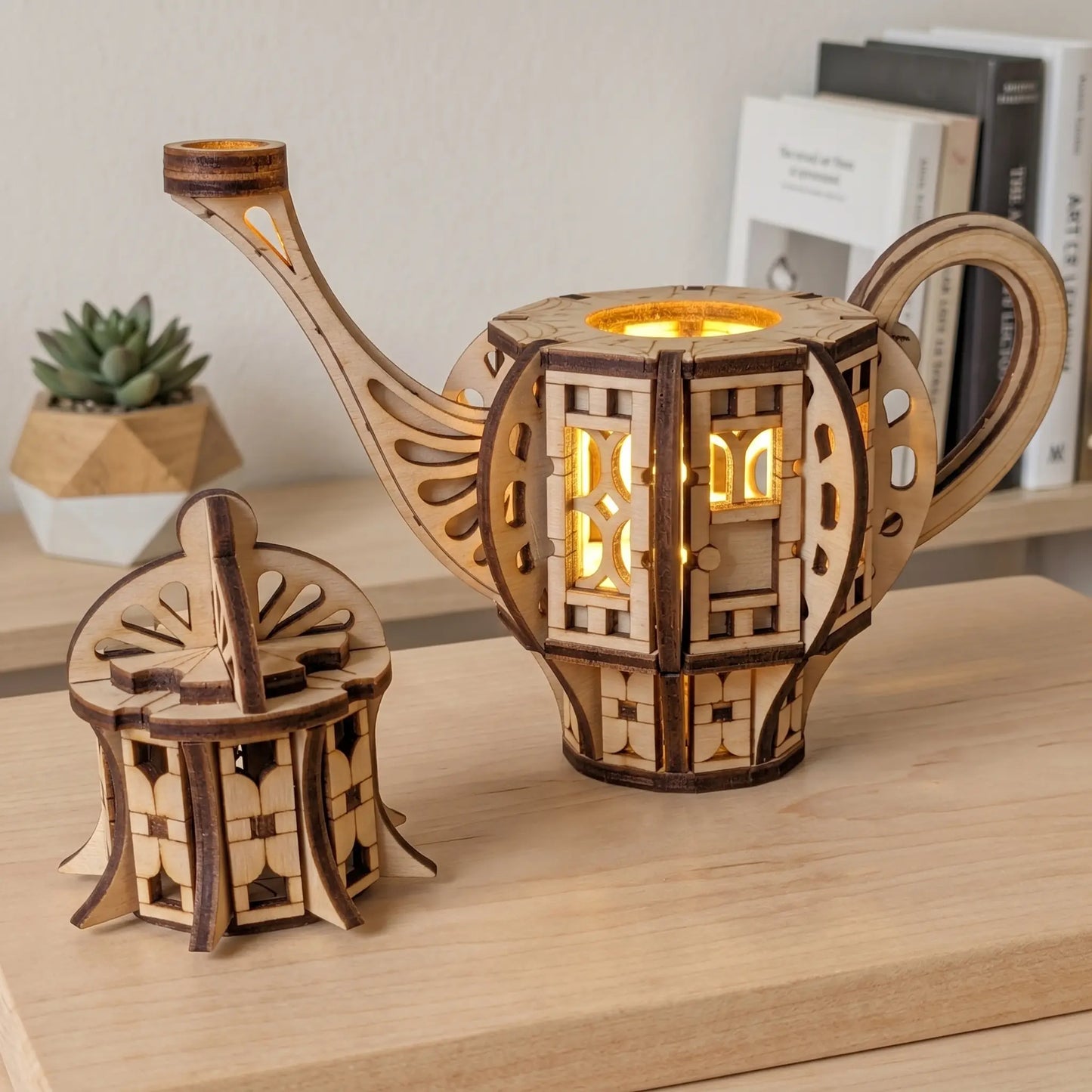Lighted teapot fairy house laser cut file with warm LED glow shining through the windows, displayed on a wooden shelf with books and a small plant.