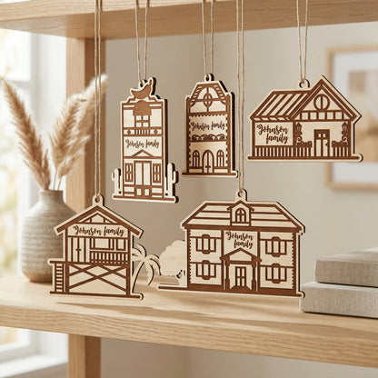 Five personalized wooden house ornaments hanging by twine strings from a shelf. The laser-cut plywood designs show various home silhouettes, each engraved with "Johnson family." Styled in a bright, modern room with minimalist decor and pampas grass.