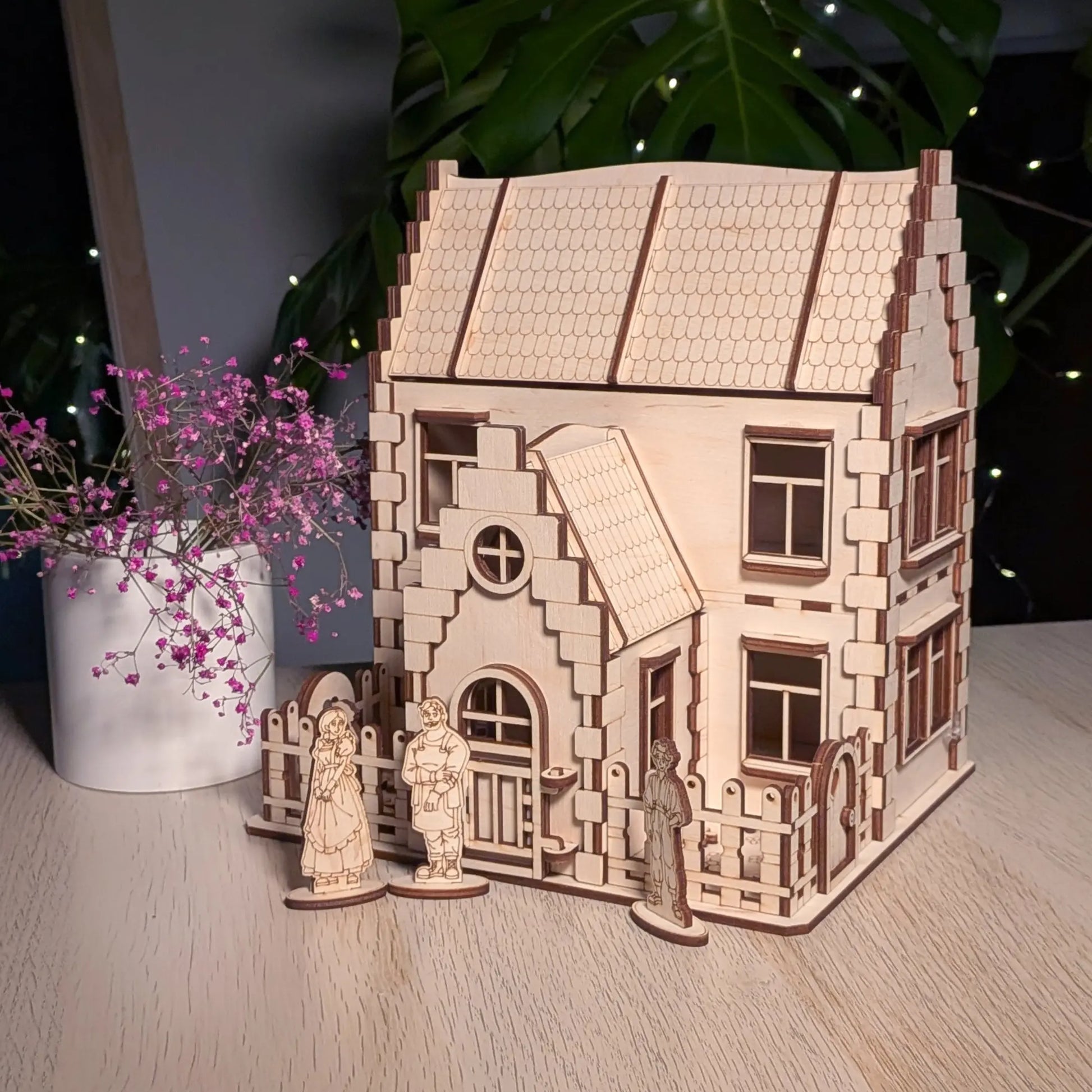 Full view of a laser cut plywood family dollhouse on a table next to a vase with small purple flowers and a green plant.
