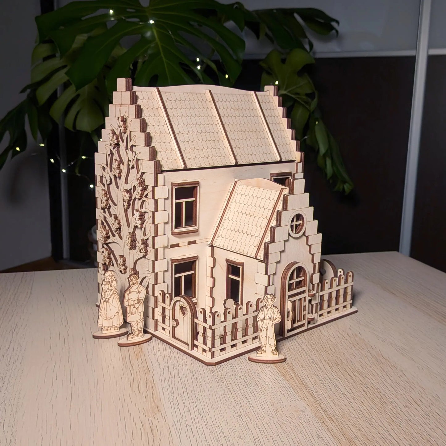 Three-quarter view of a laser cut plywood model house with three miniature wooden family figures placed around the yard.