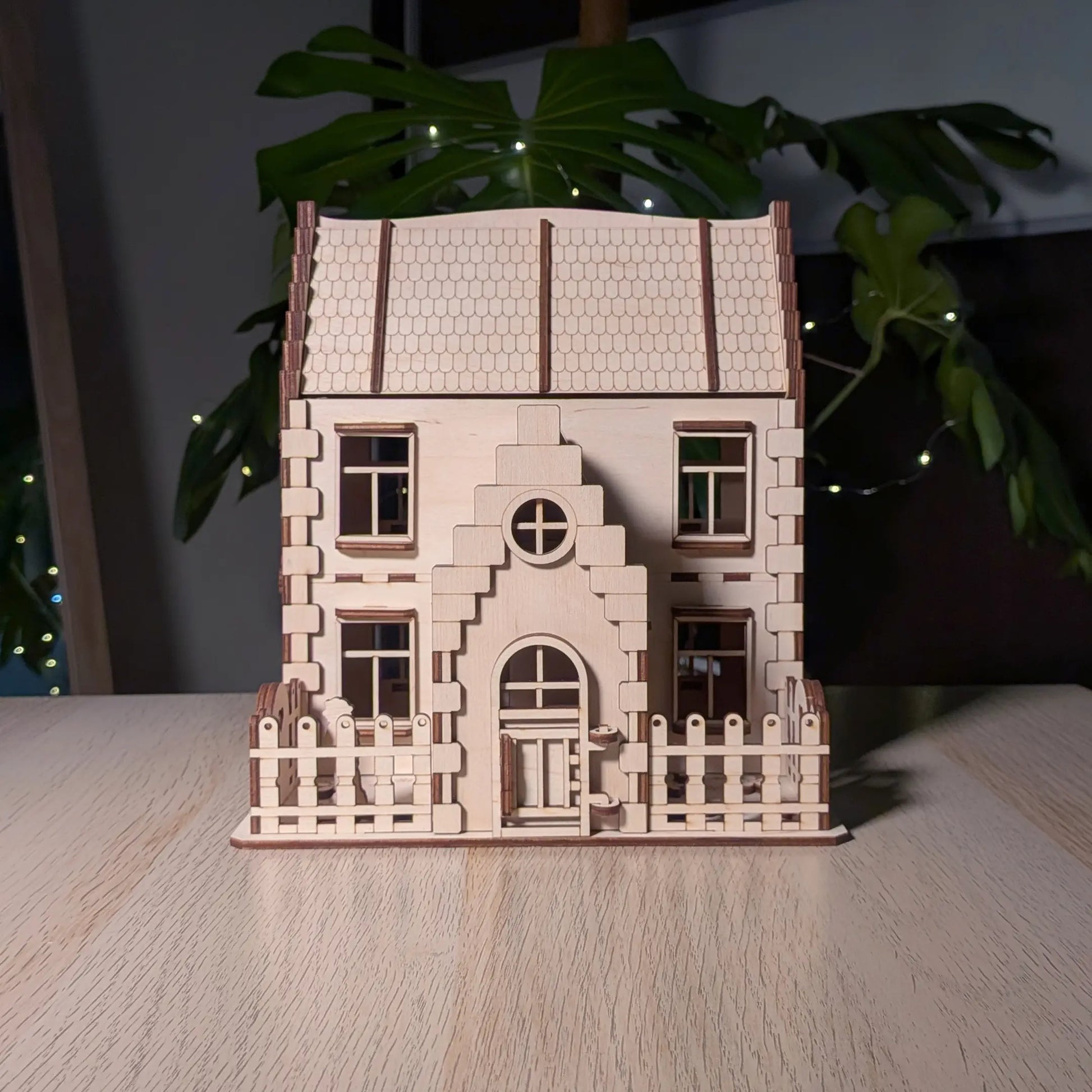 Front view of a two-story laser cut plywood family dollhouse kit featuring a small picket fence and a gabled entry door.