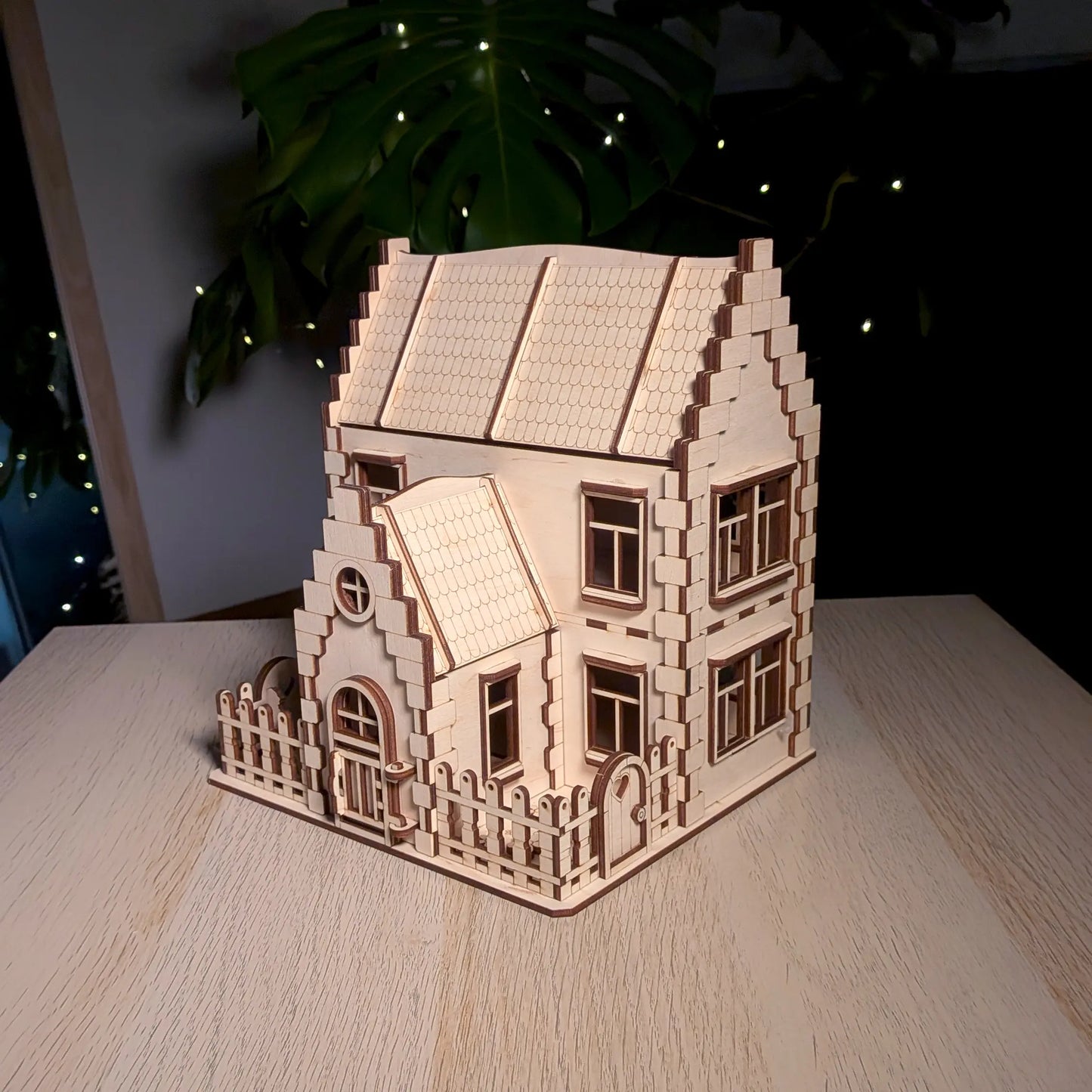 Aesthetic shot of a laser cut plywood family dollhouse on a wooden table with a large monstera plant and glowing lights.