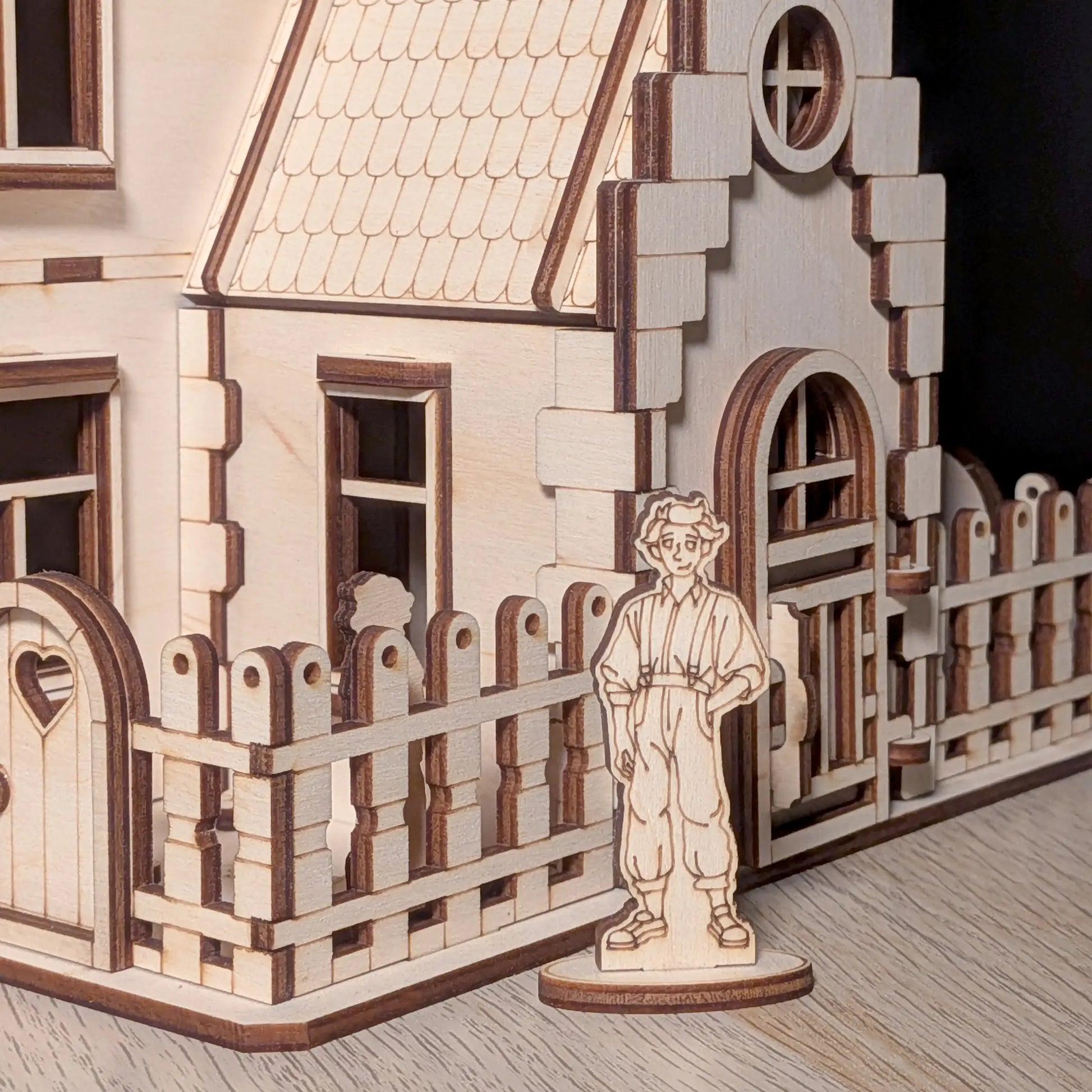 Small boy wooden figure standing next to a heart cutout gate and picket fence on a cozy laser cut plywood miniature house.