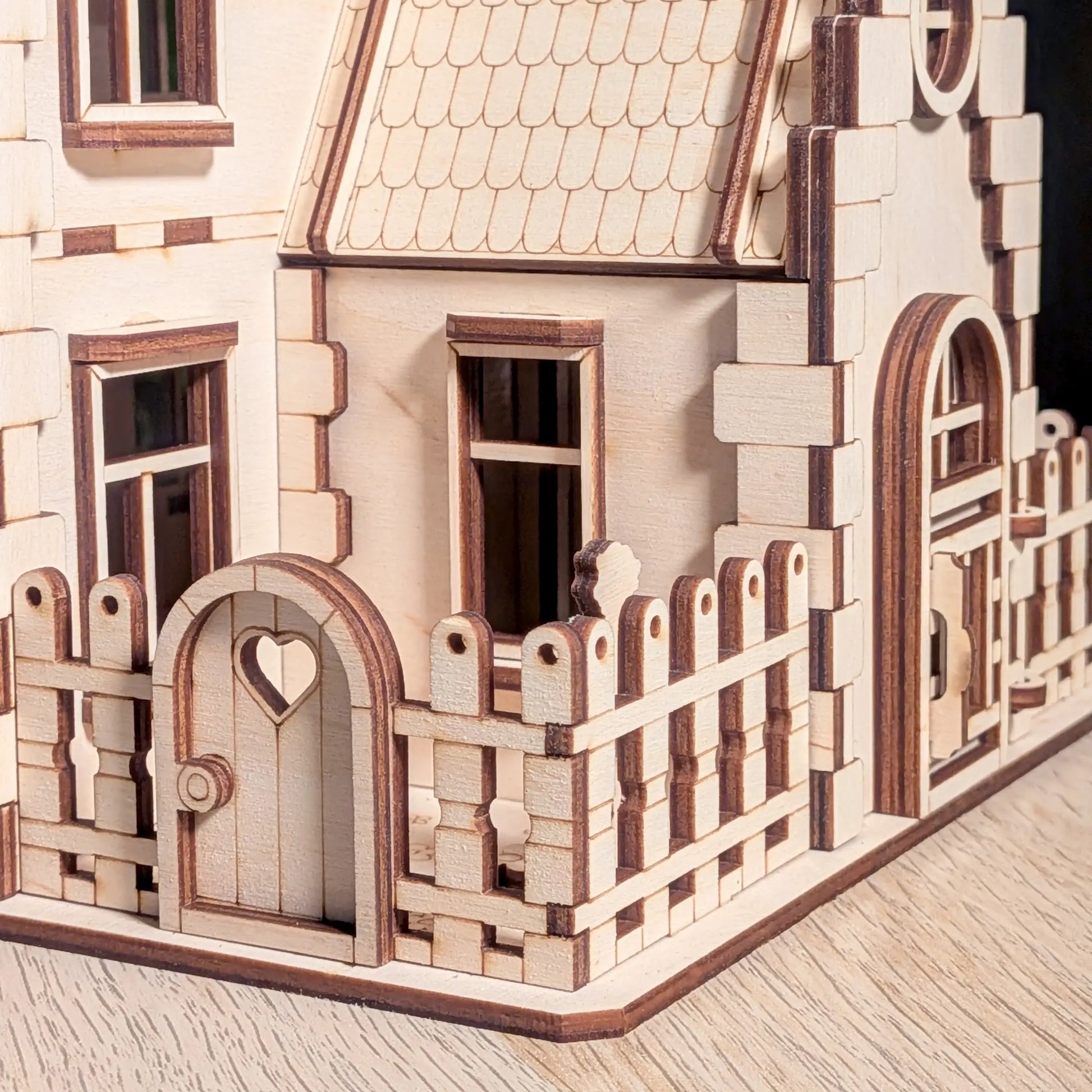 Close-up of a small picket fence with a heart cutout gate and windows on an intricate laser cut plywood family dollhouse.