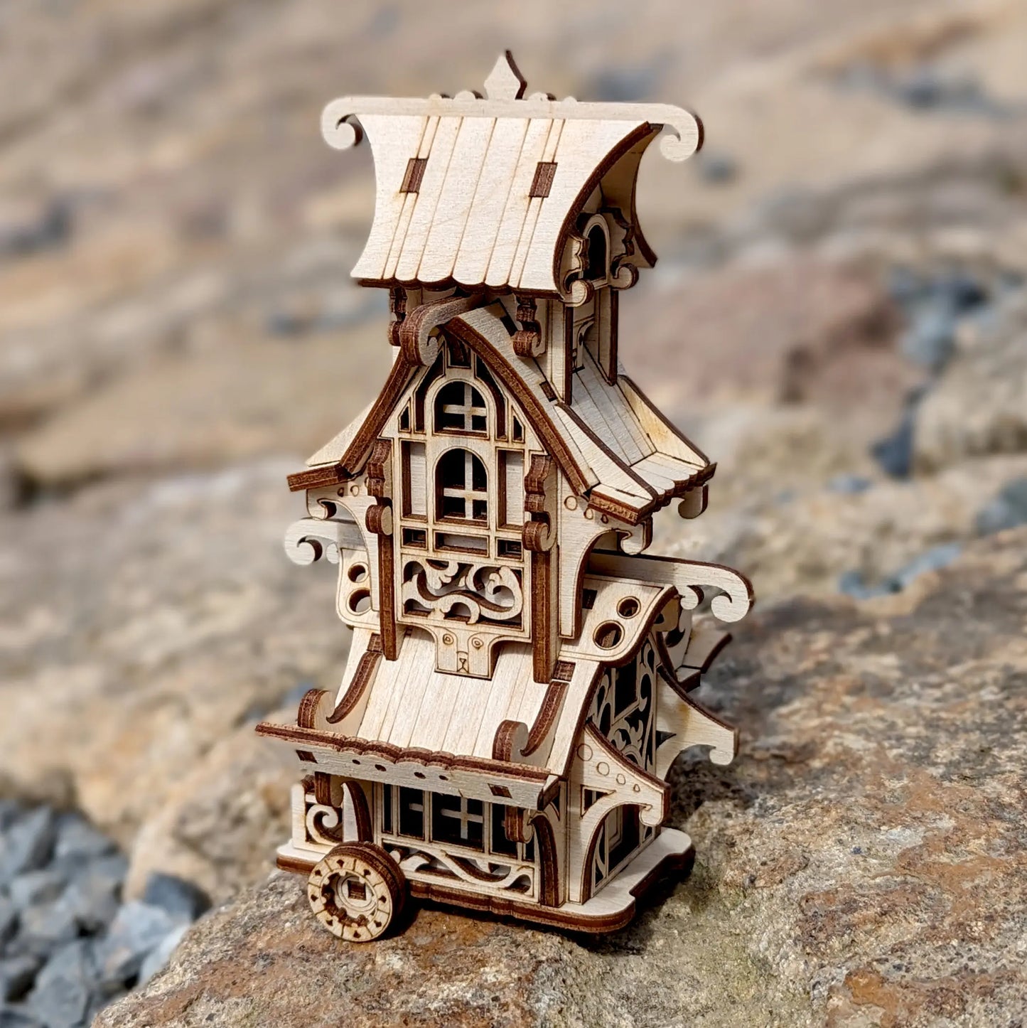 A miniature fairytale caravan model built of laser cut plywood against a blurry background of trees.