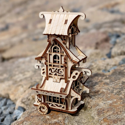 A miniature fairytale caravan model built of laser cut plywood against a blurry background of trees.