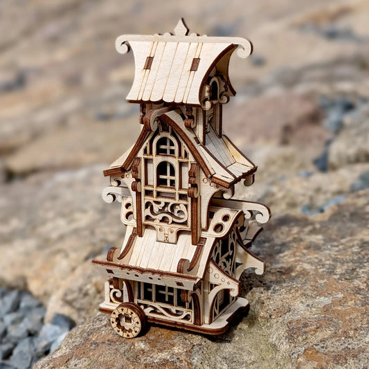 A miniature fairytale caravan model built of laser cut plywood against a blurry background of trees.