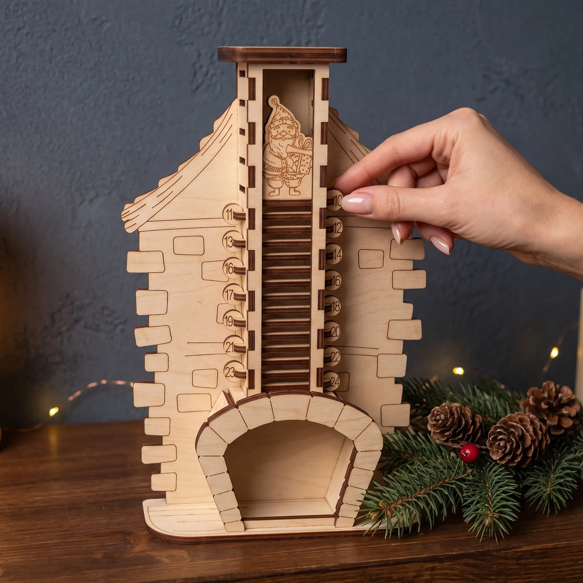 Hand removes numbered wooden slat from laser cut plywood fireplace advent calendar showing descending Santa figure