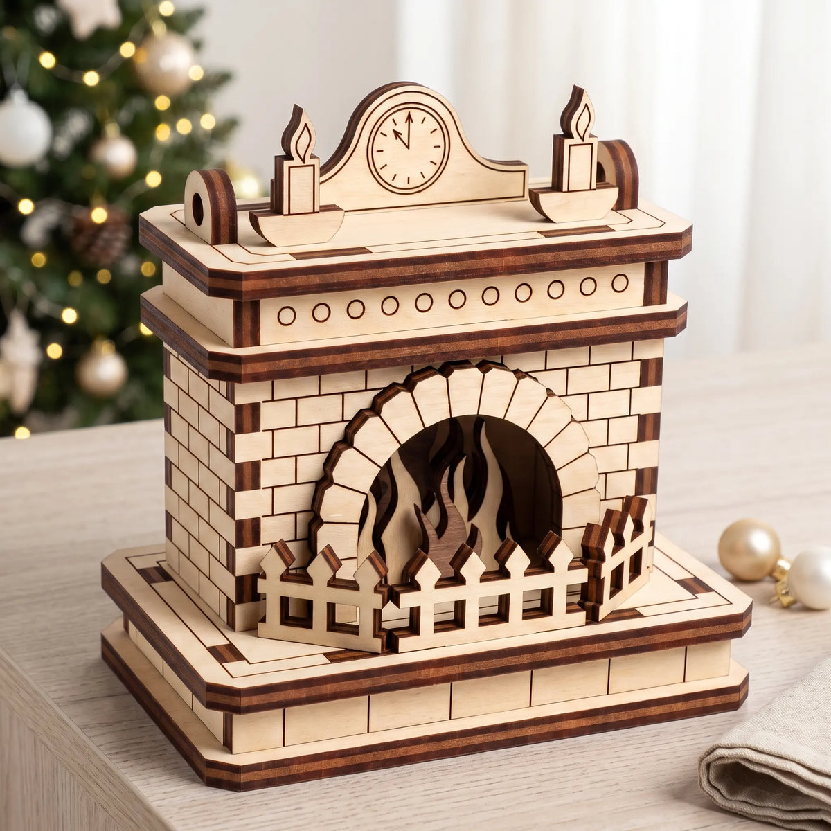Unfinished laser cut plywood fireplace model on wooden tabletop next to illuminated Christmas tree and golden ornaments.