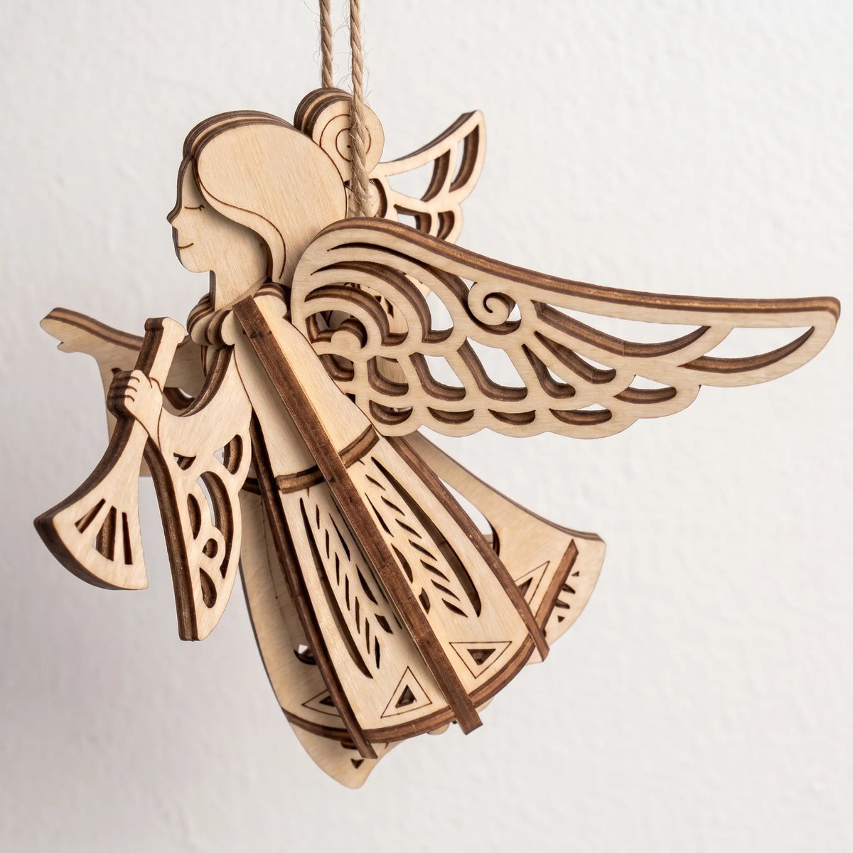 3D laser cut plywood flying angel ornament blowing a horn with carved filigree wings hanging against white wall backdrop.