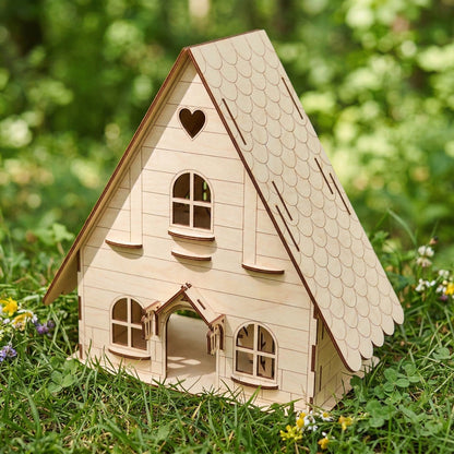Whimsical A-frame forest dollhouse made from laser cut plywood. Features a scalloped roof, heart cutout, and arched double doors. Placed on green grass in a sunny garden. Natural light wood finish with dark burnt edges.