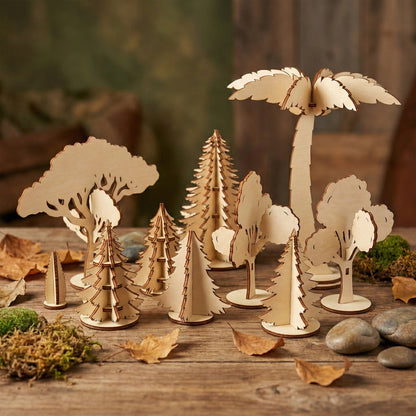 A diverse set of miniature trees made of laser cut plywood, including pines, oaks, and a tall palm tree. Arranged on a rustic wooden surface with moss, stones, and autumn leaves, showcasing the natural birch wood grain and clean edges.