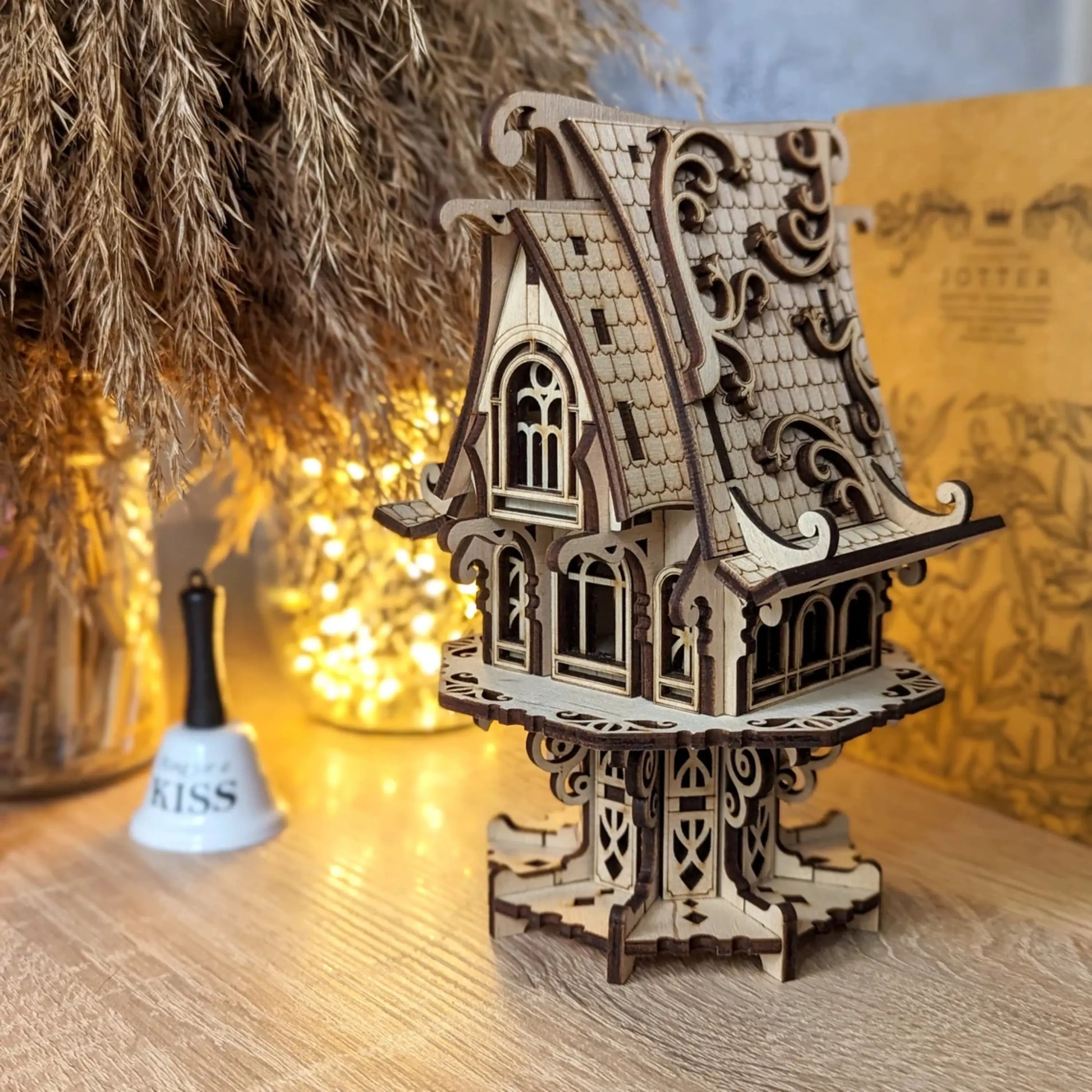 Detailed miniature elf house made of laser cut plywood stands on a desk beside a bokeh light jar and a small bell.