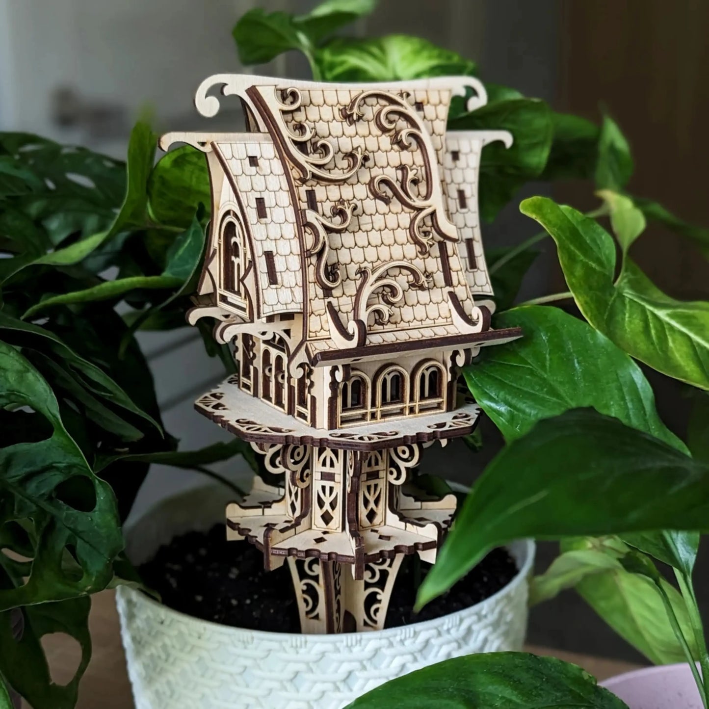 Laser cut plywood elf house stake inserted into a white textured flower pot surrounded by lush green plant leaves.
