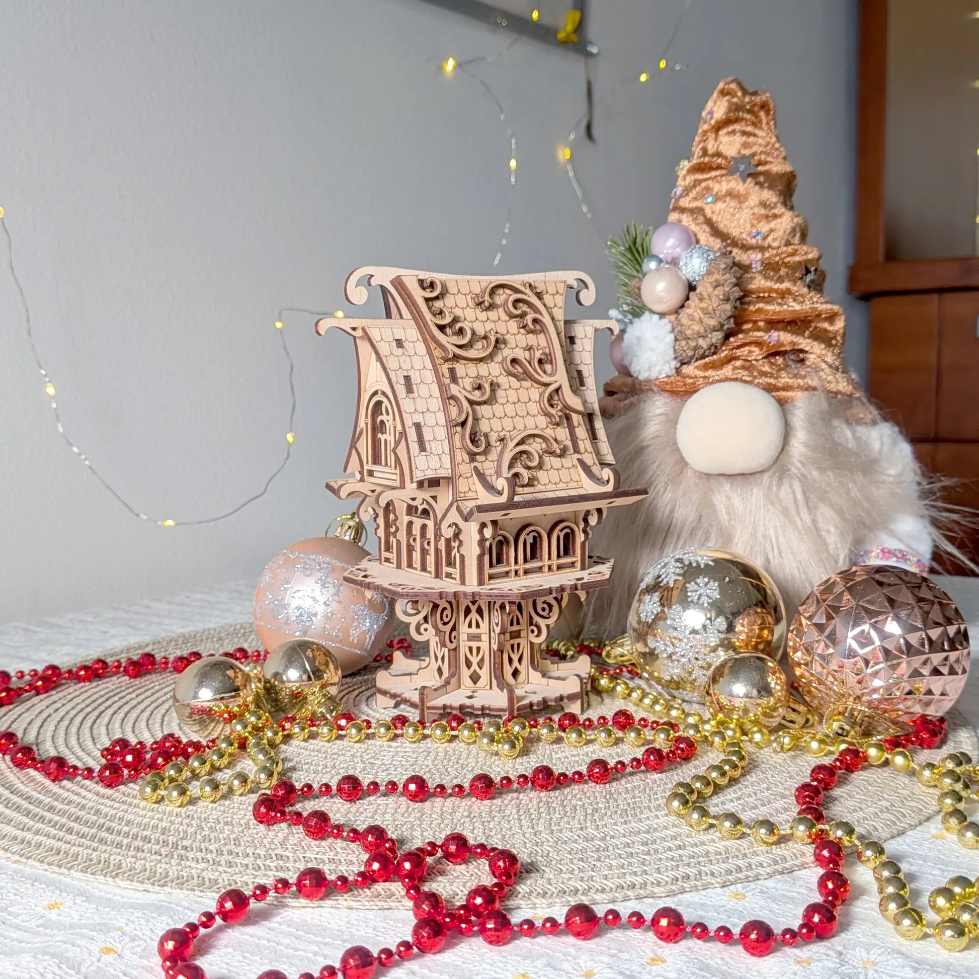 Miniature elf house of laser cut plywood sits on a woven mat surrounded by Christmas gnomes and shiny ornaments.