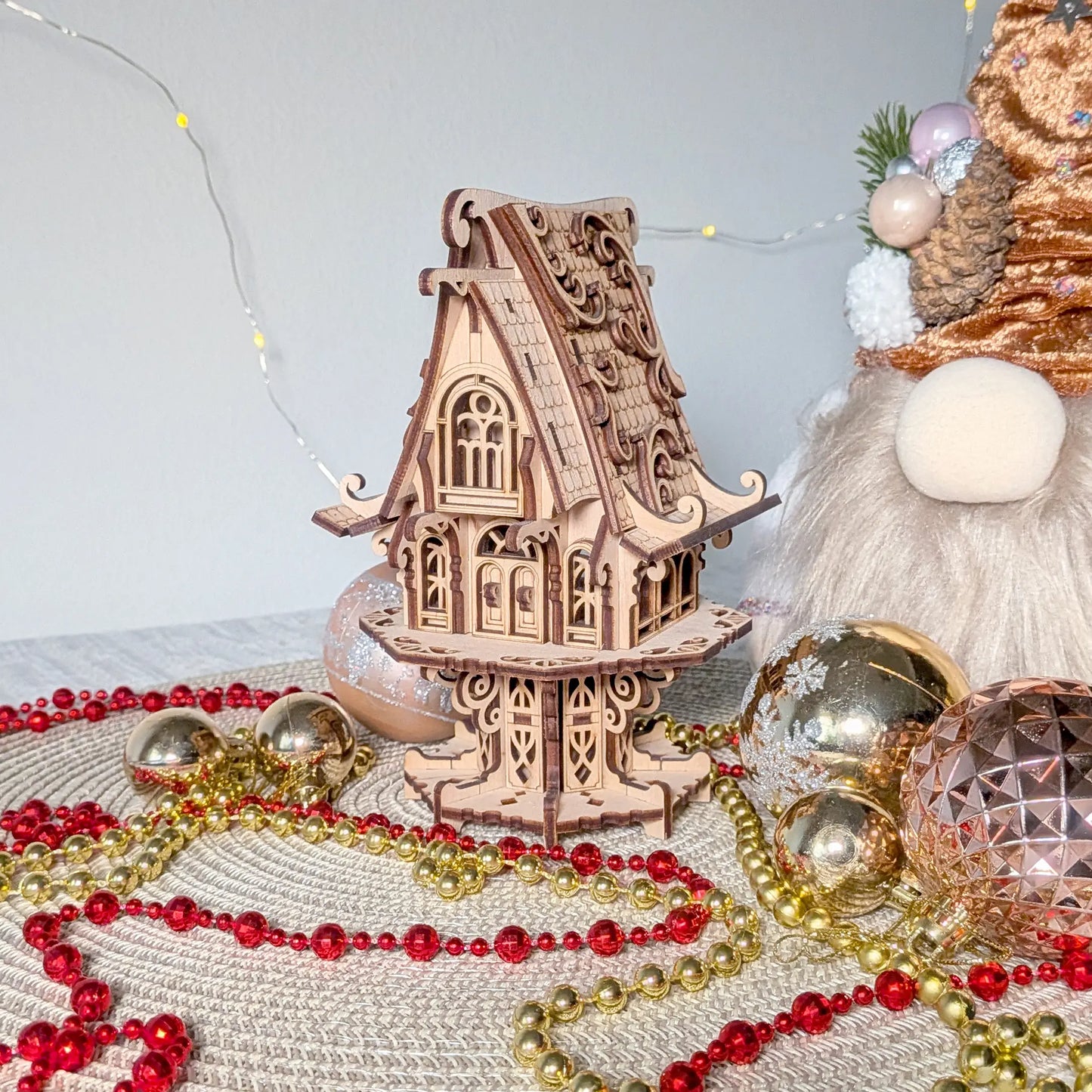 Laser cut plywood fairy elf house staged with holiday decorations, gold bead garlands, and festive glass bulbs.