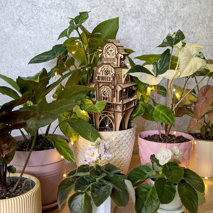 Laser cut plywood plant stake fairy castle beautifully staged amidst various potted green houseplants and lights.