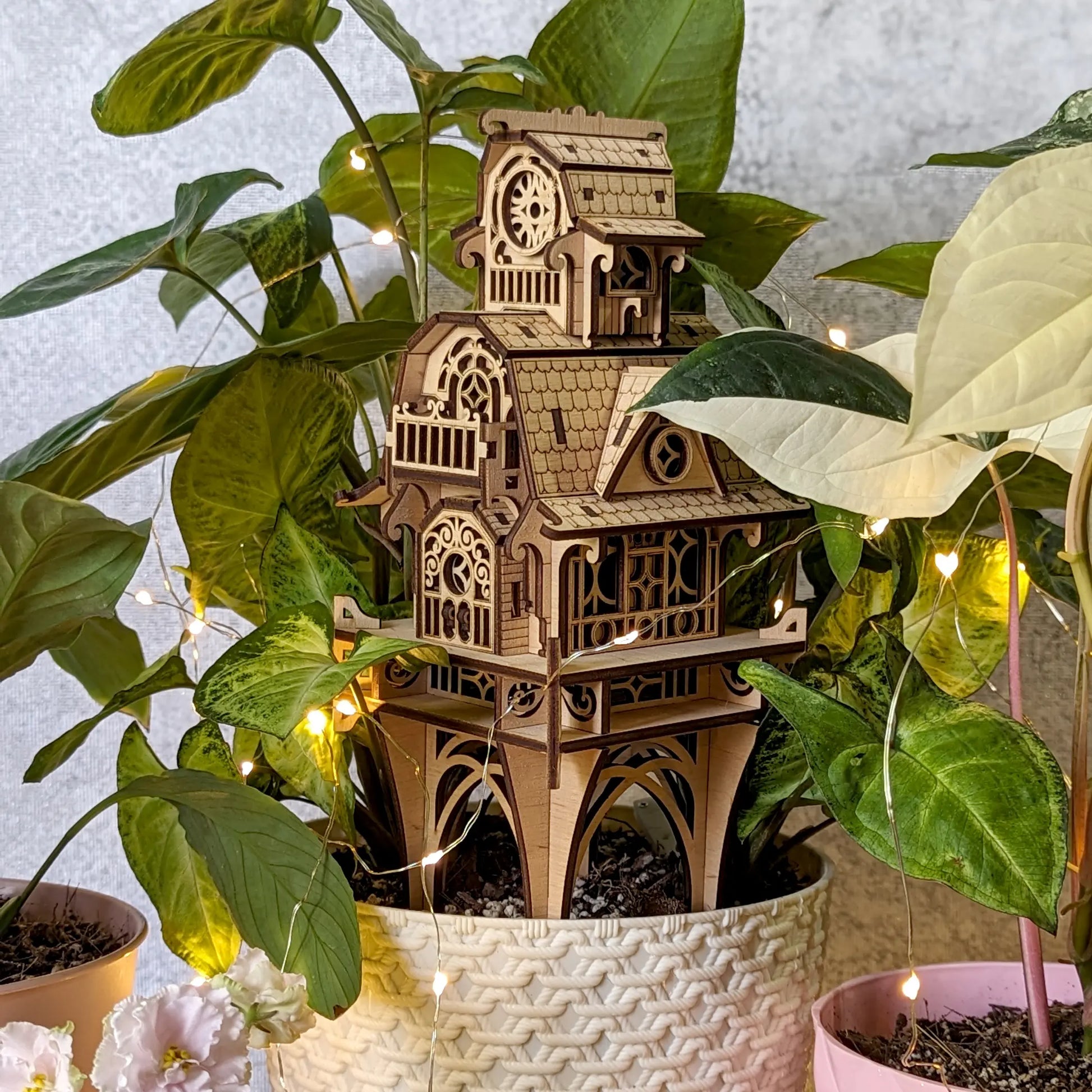 Intricate laser cut plywood garden stake house nestled into a flowerpot with delicate fairy string lights glowing.