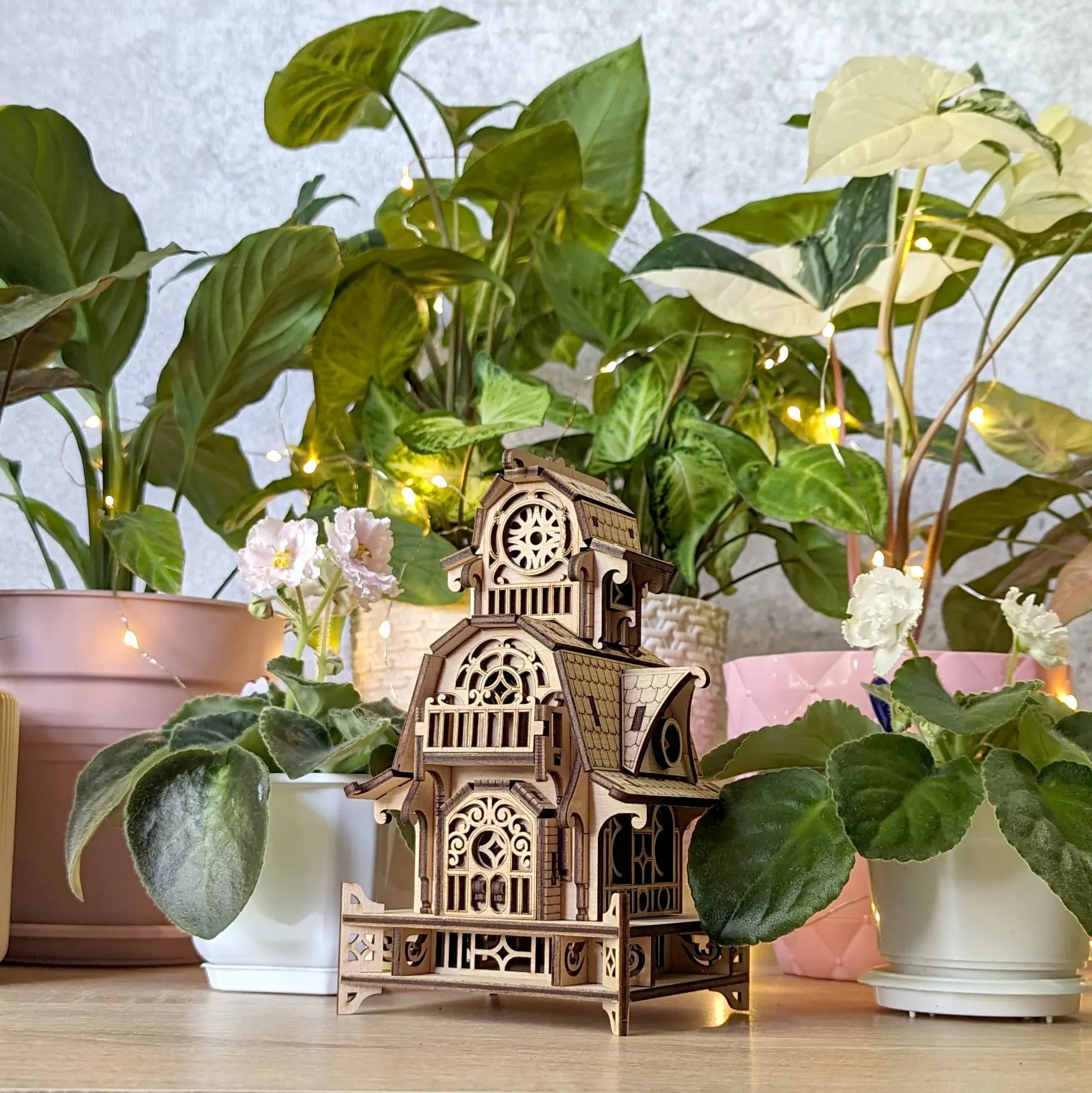 Assembled fantasy house model on a desk surrounded by indoor potted plants, showcasing its laser cut plywood frame.