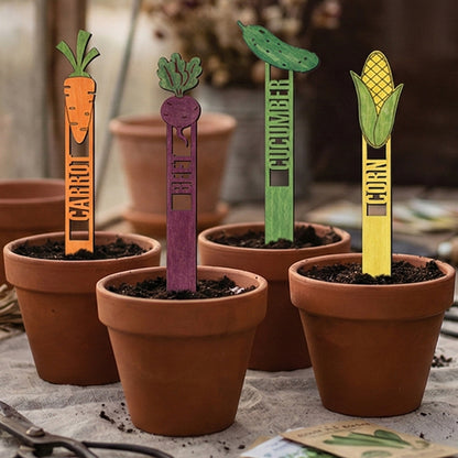 Wooden laser cut Garden Stakes 2 with vibrant designs for orange carrot, purple beet, green cucumber, and yellow corn, featuring engraved names on stakes, placed in terracotta pots against a cozy indoor gardening background.