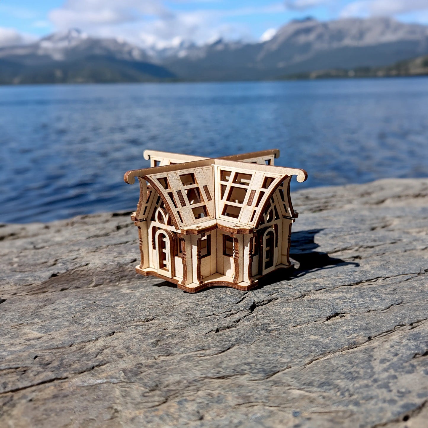 Small laser cut plywood architectural puzzle greenhouse on a riverside timber bench under a bright sky with soft clouds.